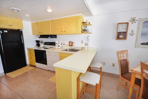 Une cuisine de taille modeste avec des placards jaunes, un réfrigérateur noir, une cuisinière et un évier. On y trouve deux chaises en bois et une petite table.