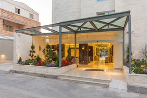 Entrada de hotel con fachada de vidrio, zona exterior cubierta y jardín ajardinado con diversas plantas.