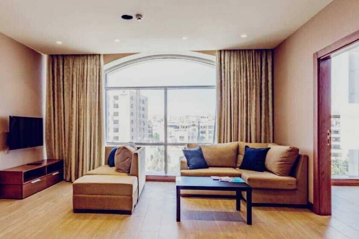 Una sala moderna con sofá, reposapiés y mesa de centro, ventanas grandes con cortinas y un televisor de pantalla plana sobre un mueble de madera.