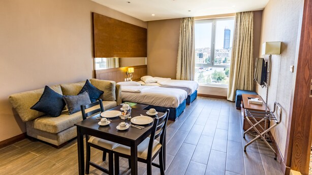 A hotel room with a sofa, a dining table set with plates and cutlery, a bed, a window with curtains, and a lamp on the wall. - The Seventh Star Hotel Suites (Amman)
