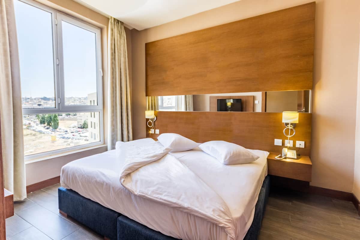 Habitación de hotel con una cama grande, cabecera de madera, dos lámparas de noche, mesita de noche y vistas de la ciudad a través de un ventanal.