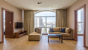 A modern living room with a sofa, ottoman, and coffee table, large windows with curtains, and a flat-screen TV on a wooden stand. - The Seventh Star Hotel Suites (Amman)