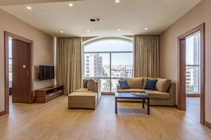 Una sala de estar moderna con sofá, reposapiés y mesa de centro, ventanas amplias con cortinas y un televisor de pantalla plana sobre un mueble de madera.