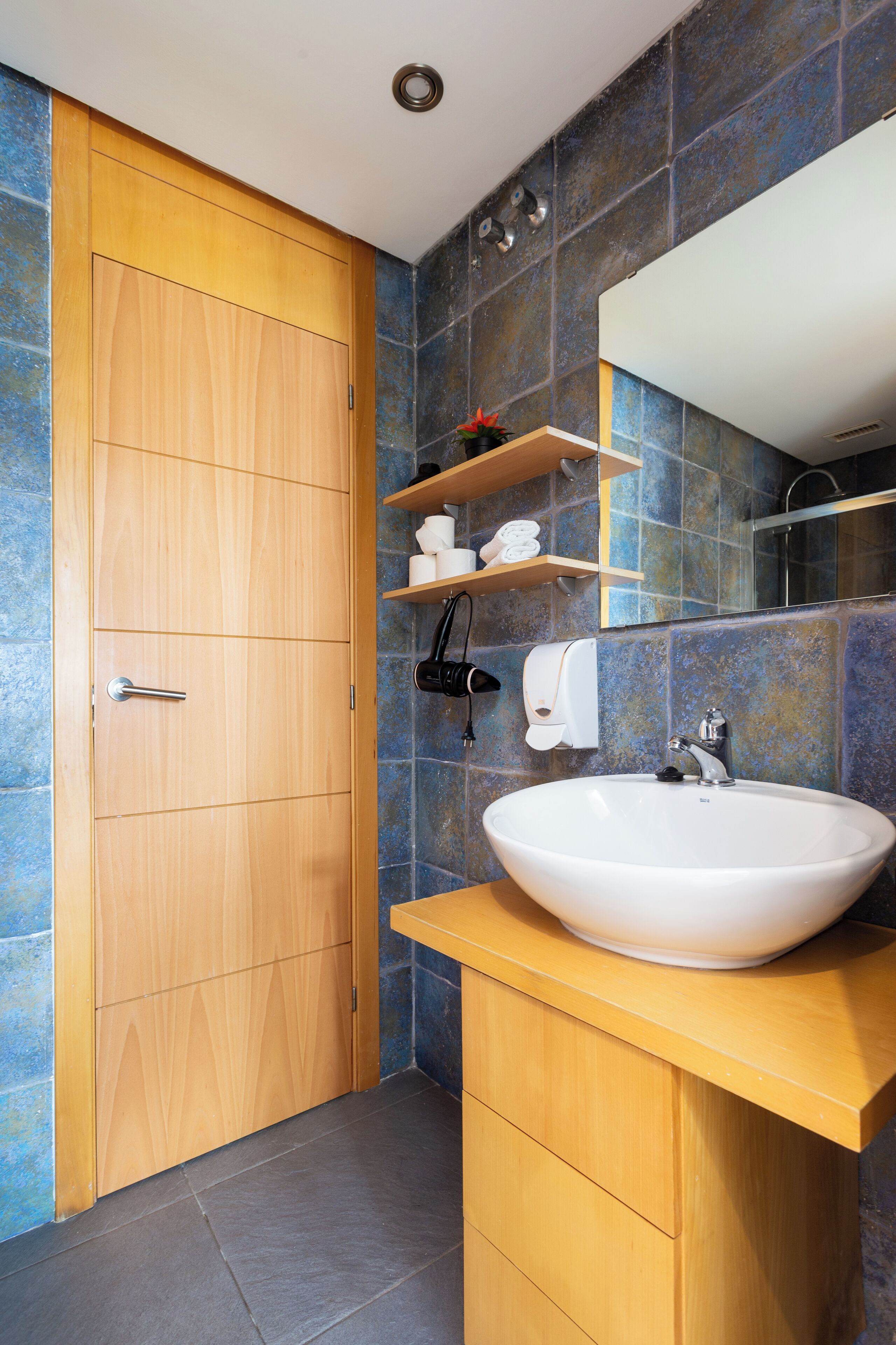A bathroom with a wooden door, a white sink, a mirror, and a shelf with toiletries.