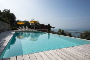A rectangular swimming pool with clear blue water, surrounded by a wooden deck. There are two yellow umbrellas providing shade, and the pool is adjacent to a building with a view of the sea. - Gianni with 3 bedrooms with balcony and lake view (Torri del Benaco)