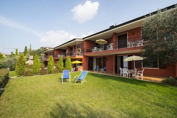 A two-story building with a red facade, outdoor seating area with blue and white chairs, and a small garden. - Gianni with 3 bedrooms with balcony and lake view (Torri del Benaco)