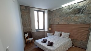 Un dormitorio con pared de piedra, cama de madera, mesita de noche, una silla y una ventana.