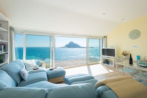 A modern living room with a large window overlooking the sea, a comfortable seating area, a flat-screen TV, and a dining table with chairs. - Cockleshell Cottage, pristine luxury and tranquility (Marazion)
