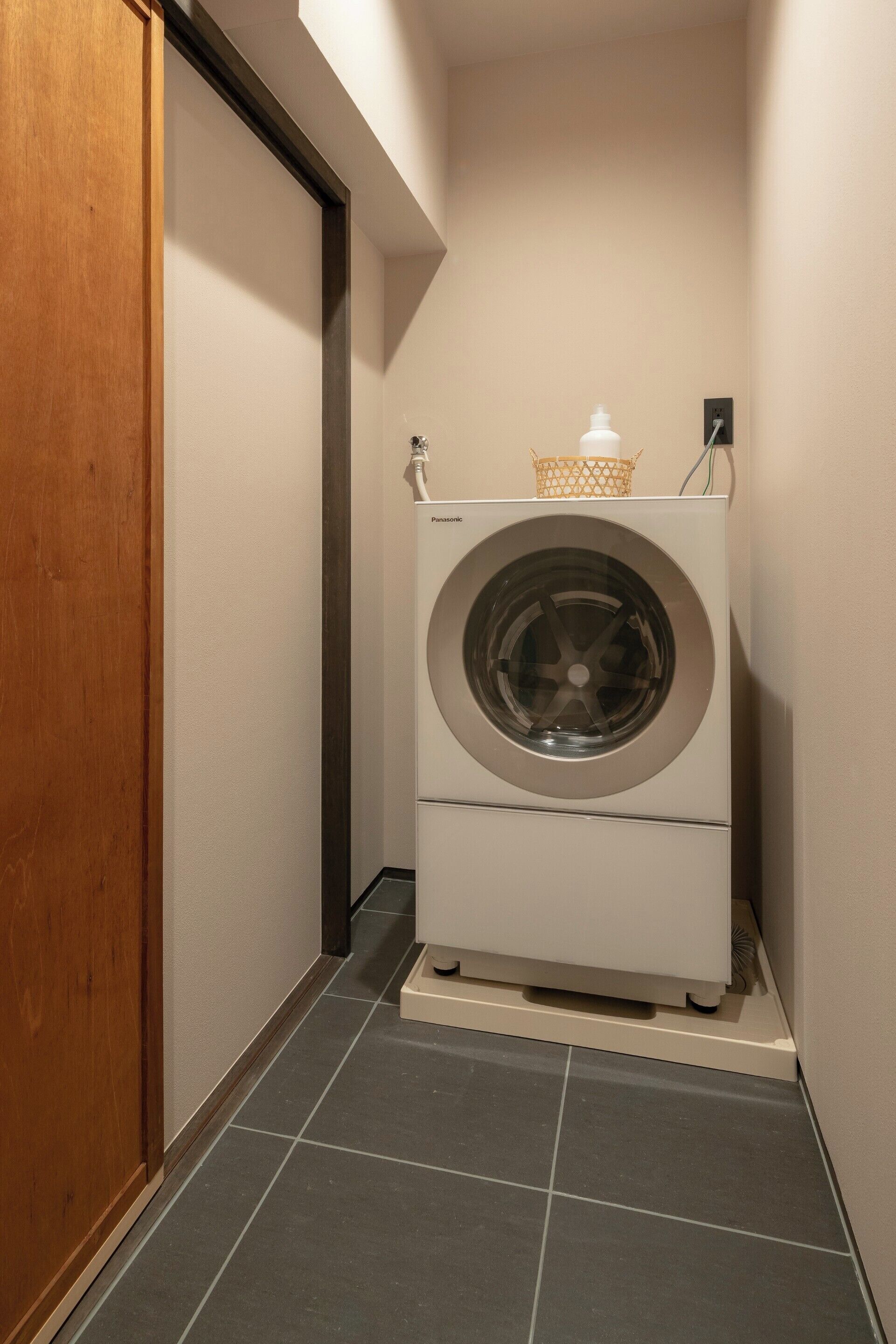 A compact laundry room with a front-loading washer and dryer, a wooden door, and a tiled floor.