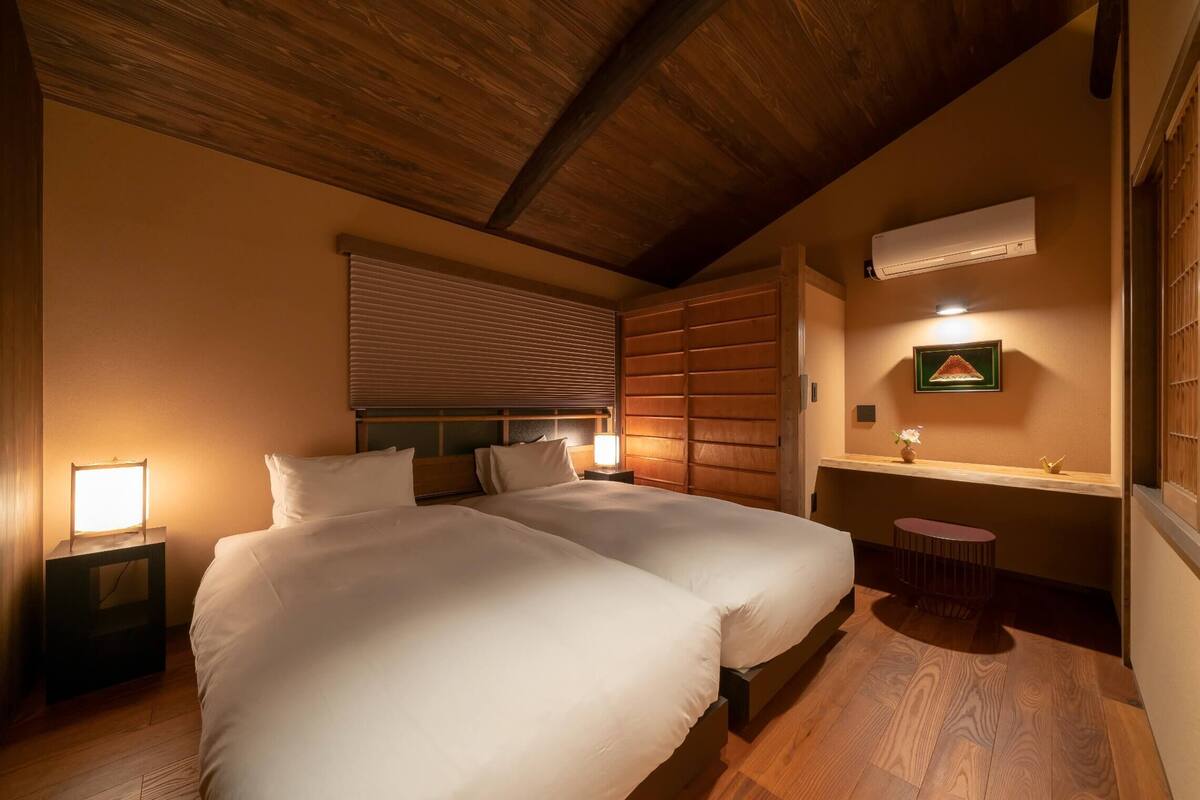 A bedroom with two beds, a bedside table with a lamp, a window with blinds, a wall-mounted air conditioner, and a small table with a plant.