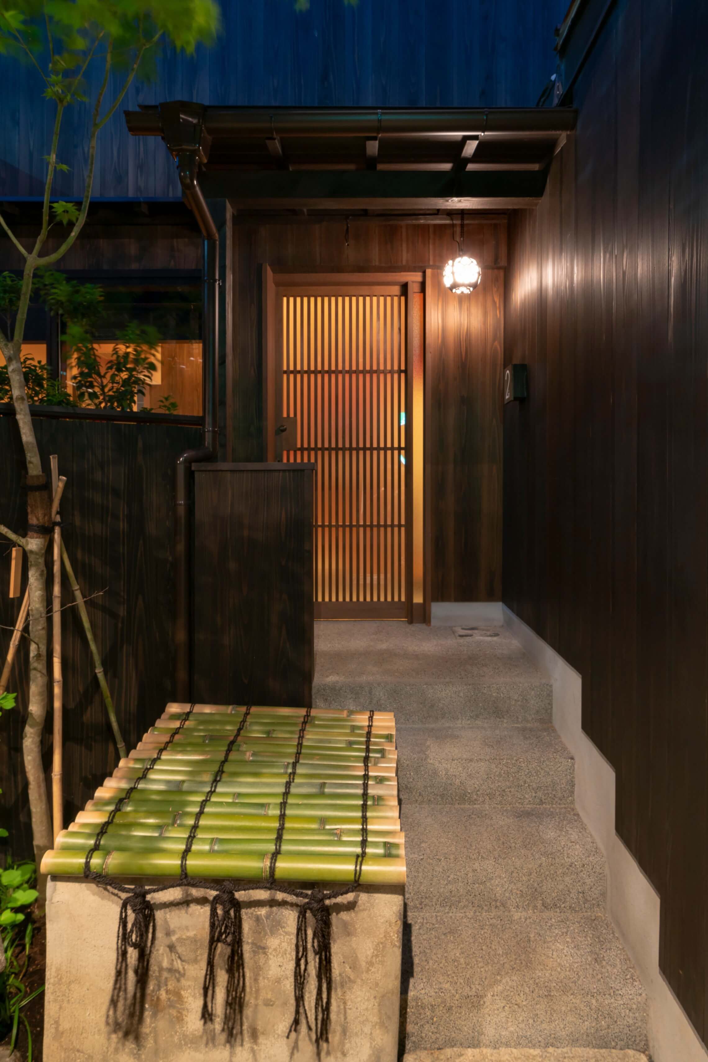 A traditional Japanese-style entrance with a wooden gate, a stone lantern, and a concrete step leading to a wooden door.