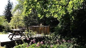 A garden patio with a wooden deck, surrounded by lush greenery and flowering plants. - Lily cottage In An Idyllic Riverside Location, Lake District Nation Park. (Selside Kendal,)
