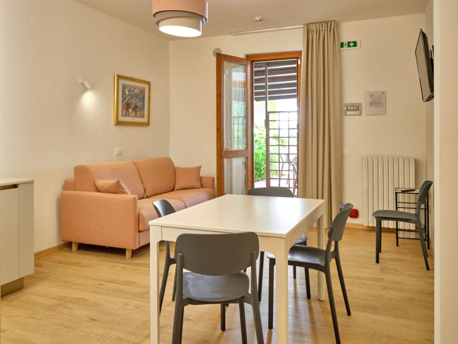 Una habitación moderna con piso de madera, una mesa blanca con cuatro sillas, un sofá color melocotón, una ventana con persianas y una señal de salida verde.