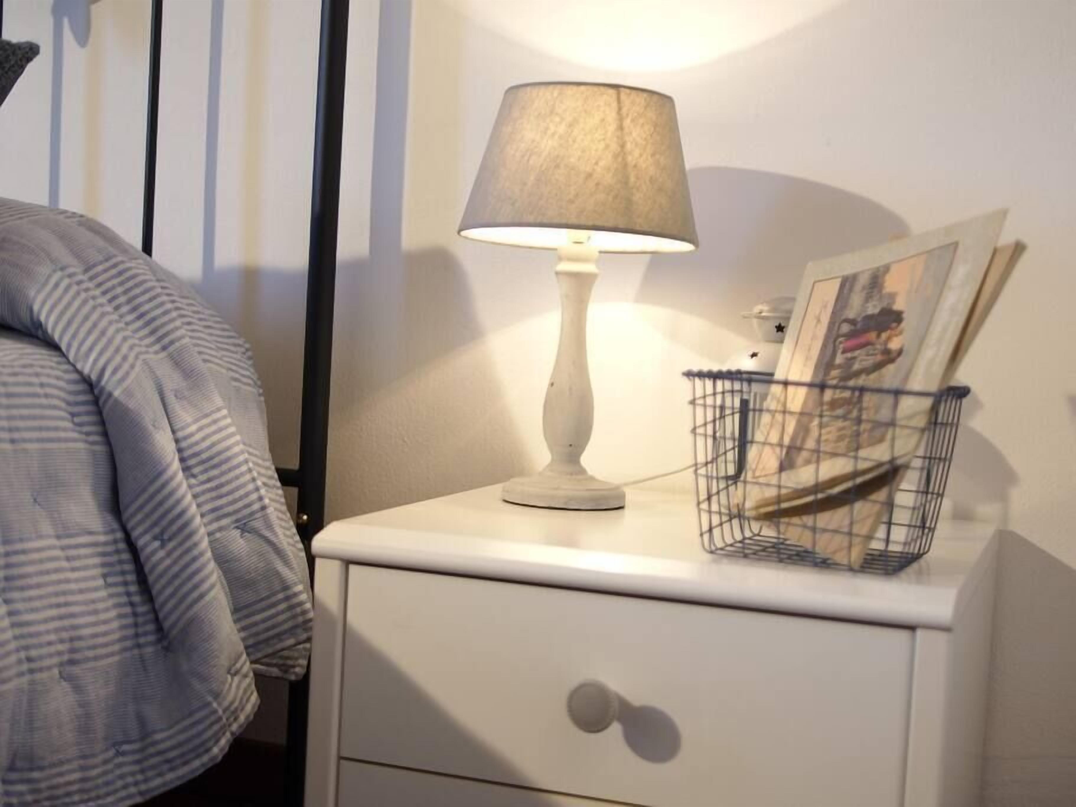 Une table de chevet avec une lampe, un petit panier en fil de fer et un tableau encadré.