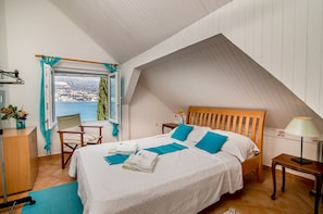A bedroom with a sloped ceiling, a bed with blue bedding, a wooden headboard, a nightstand, a lamp, a chair, a desk, and a window with a view of a snowy landscape. - Beautiful shoreline holiday home with private mooring, Njivice, Montenegro (Njivice)
