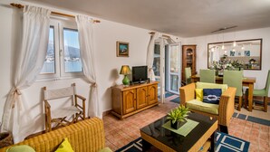 A living room with a television, wicker furniture, a dining area with chairs and a table, and a view of a body of water through the windows. - Beautiful shoreline holiday home with private mooring, Njivice, Montenegro (Njivice)