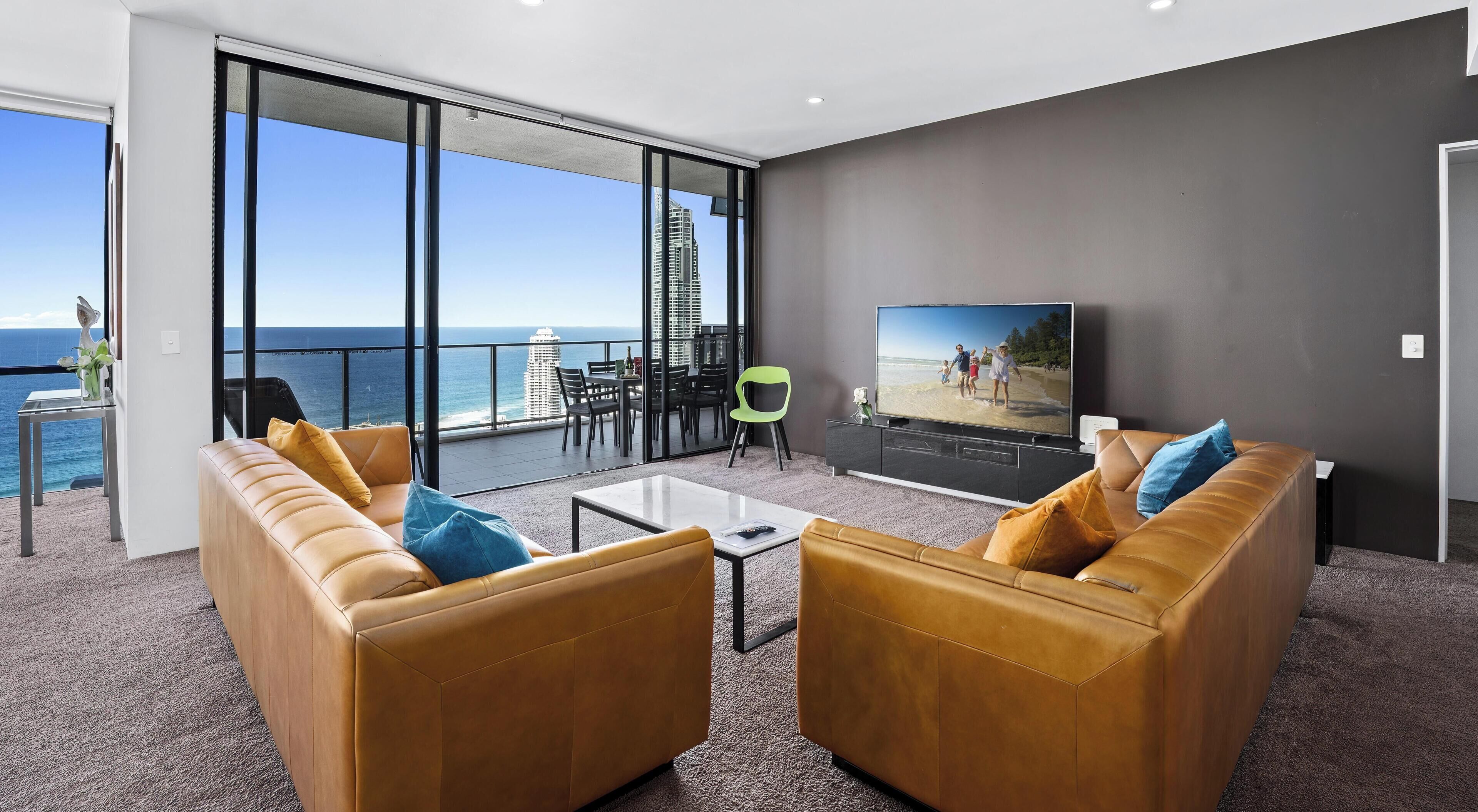 A modern living room with a large TV, leather sofas, a glass table, and a balcony with a view of the ocean and buildings.