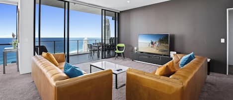 A modern living room with a large TV, leather sofas, a glass table, and a balcony with a view of the ocean and buildings.