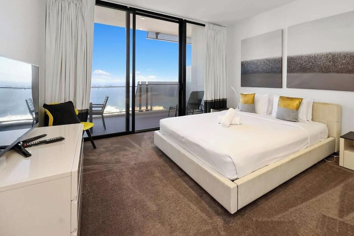 A hotel room with a bed, a desk, a chair, a nightstand, and a view of the sea.