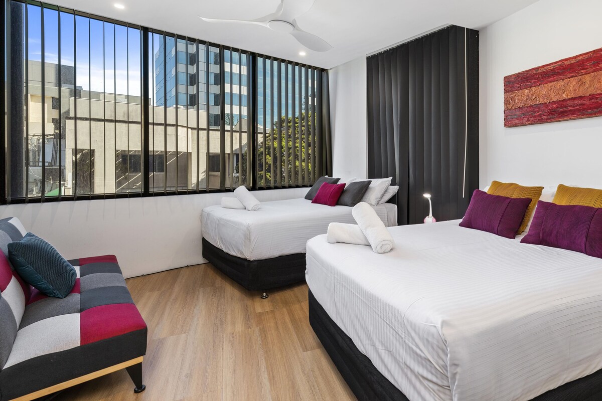 A modern hotel room with two beds, a sofa, and a large window with blinds.