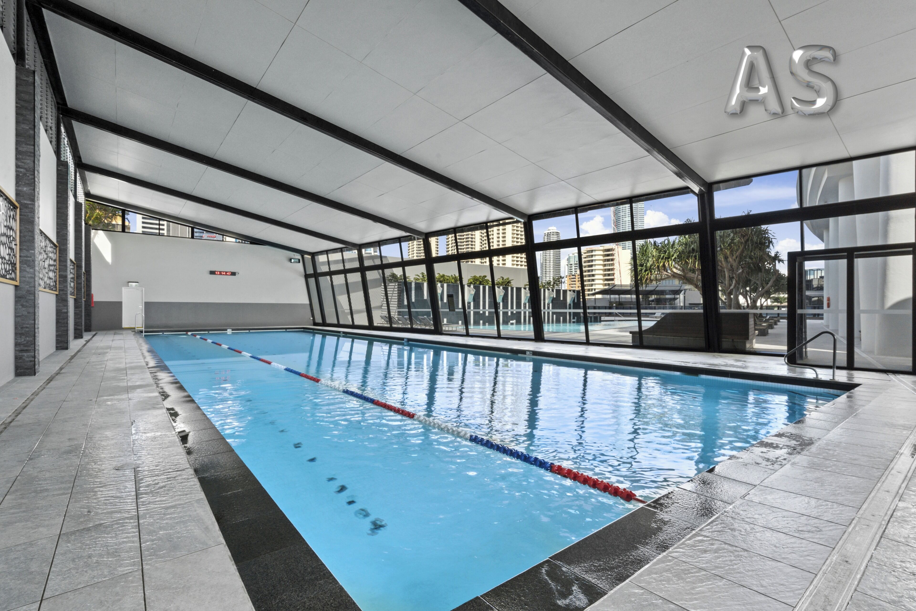 An indoor swimming pool with clear water, black tiled flooring, and large windows offering views of the city.