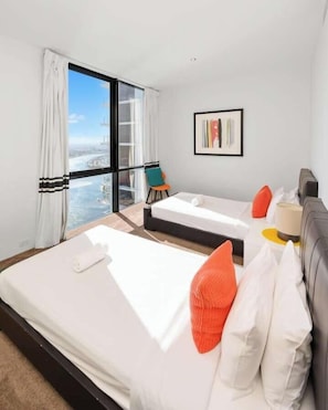 A modern hotel room with two beds, a view of the city, a chair, and a framed artwork.