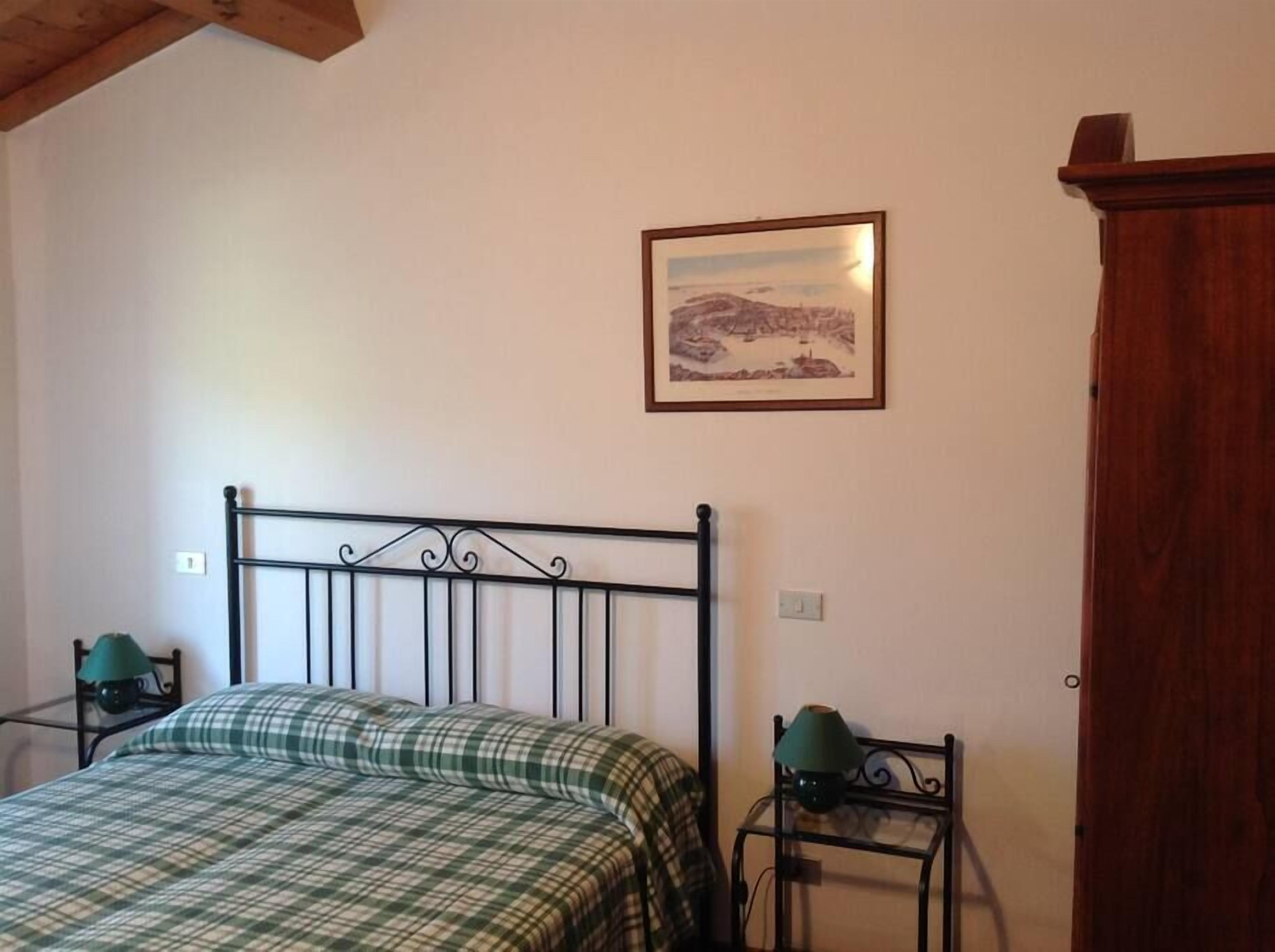 A bedroom with a metal bed frame, a green plaid bedspread, a framed landscape painting on the wall, and a wooden cabinet.