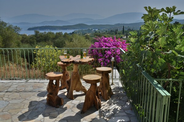 Una terraza con mesas y sillas de madera, rodeada de follaje y con vistas a un lago y montañas.