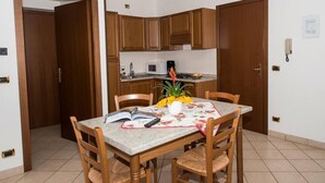 A dining area with a table set for two, wooden chairs, a book, a vase with flowers, and a microwave in the background.