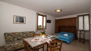 A compact room with a bed, a dining table with chairs, a patterned sofa, and a small kitchenette area.