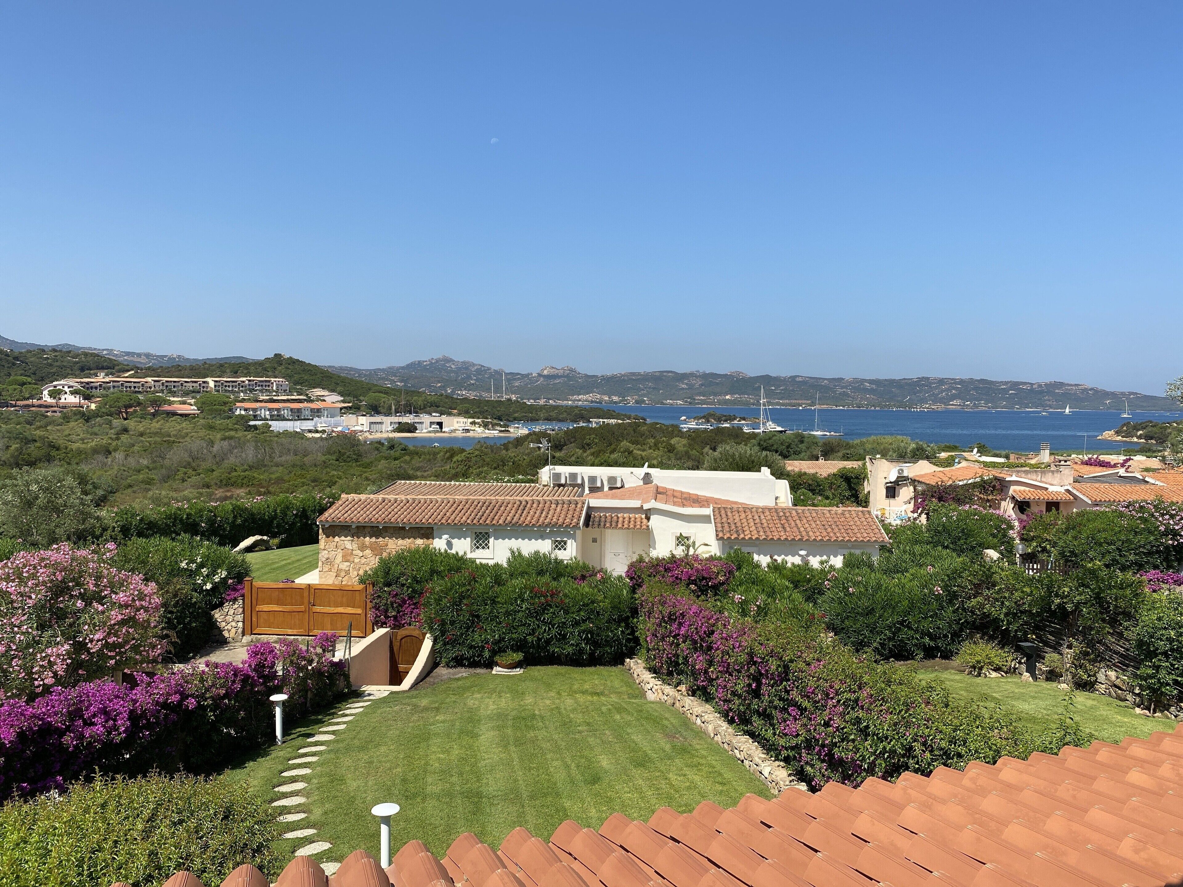 Una villa costera con un jardín cuidado, un sendero de piedra y vistas al mar y barcos a lo lejos.