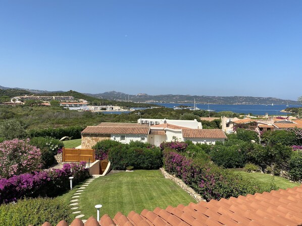 A coastal villa with a well-maintained garden, a stone pathway, and a view of the sea and boats in the distance.