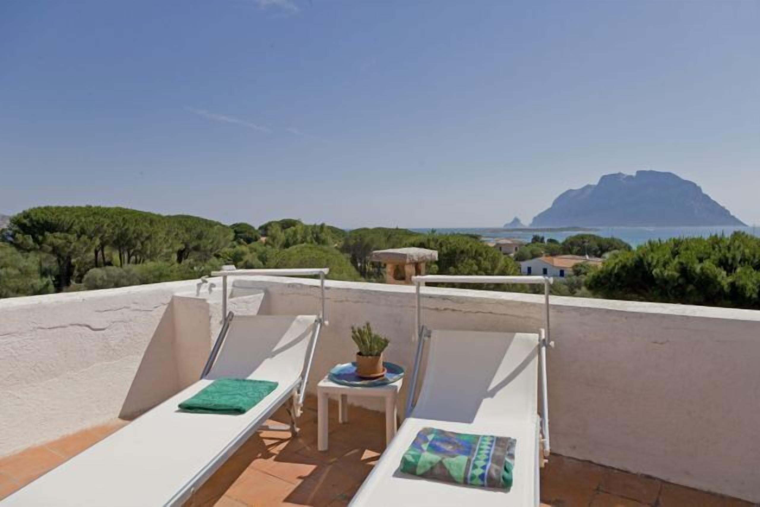 Een dakterras met twee ligbedden, een kleine tafel met een plant in pot, en uitzicht op een berg en zee.