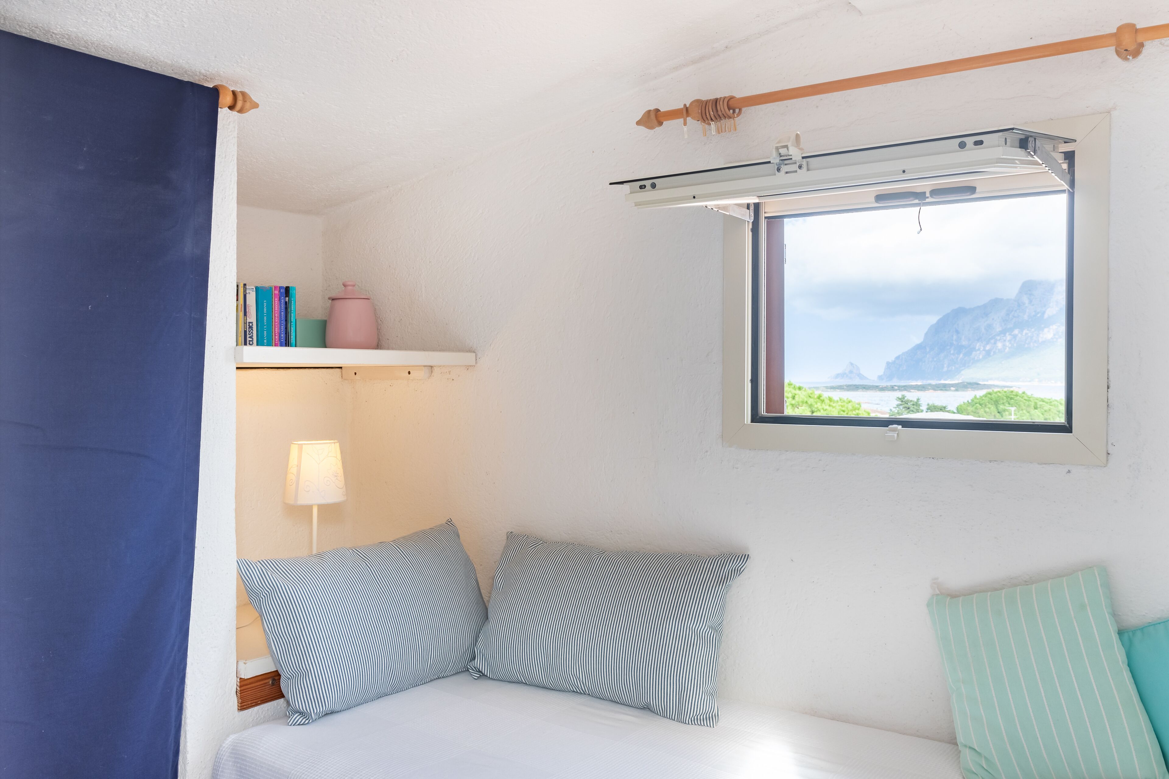 Un dormitorio con cama, almohadas, una estantería con libros y un recipiente rosa, una ventana con vista a acantilados y un ventilador de techo.