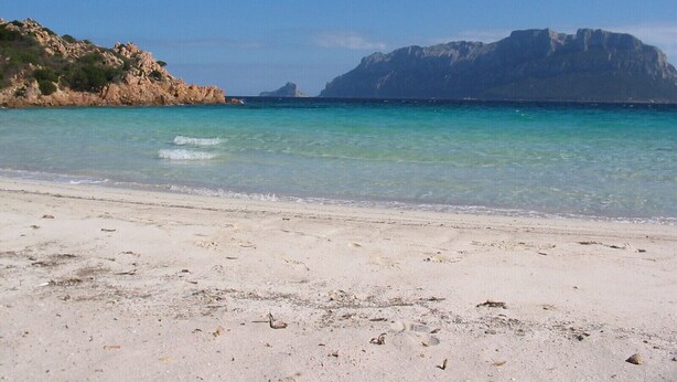 A beach with clear turquoise water, sandy shore, and rocky cliffs in the background.