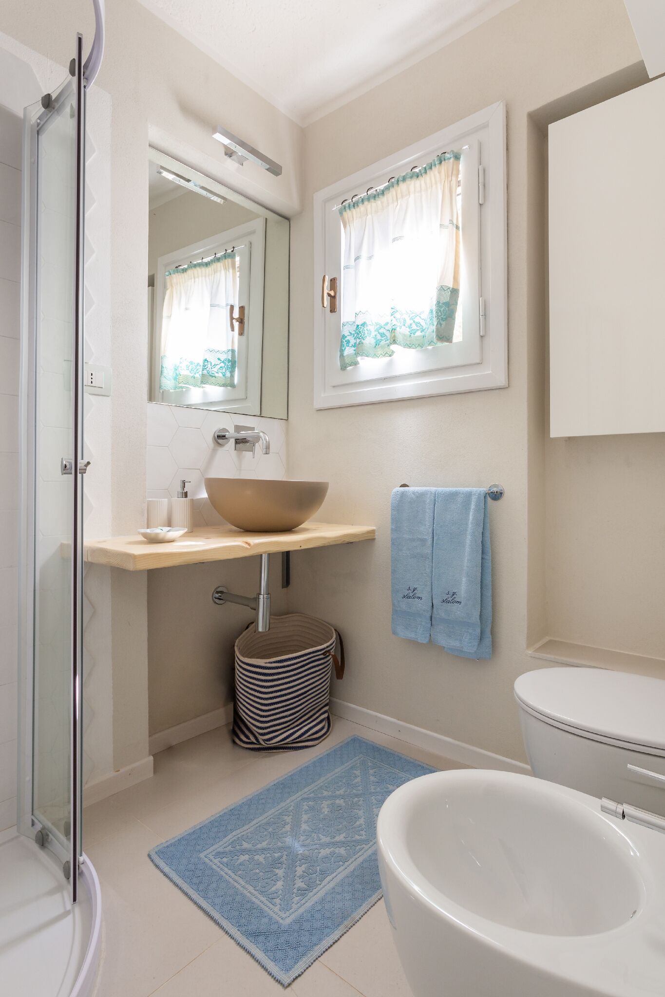 A bathroom with a glass shower enclosure, a beige sink with a large bowl, a blue towel, and a white toilet.