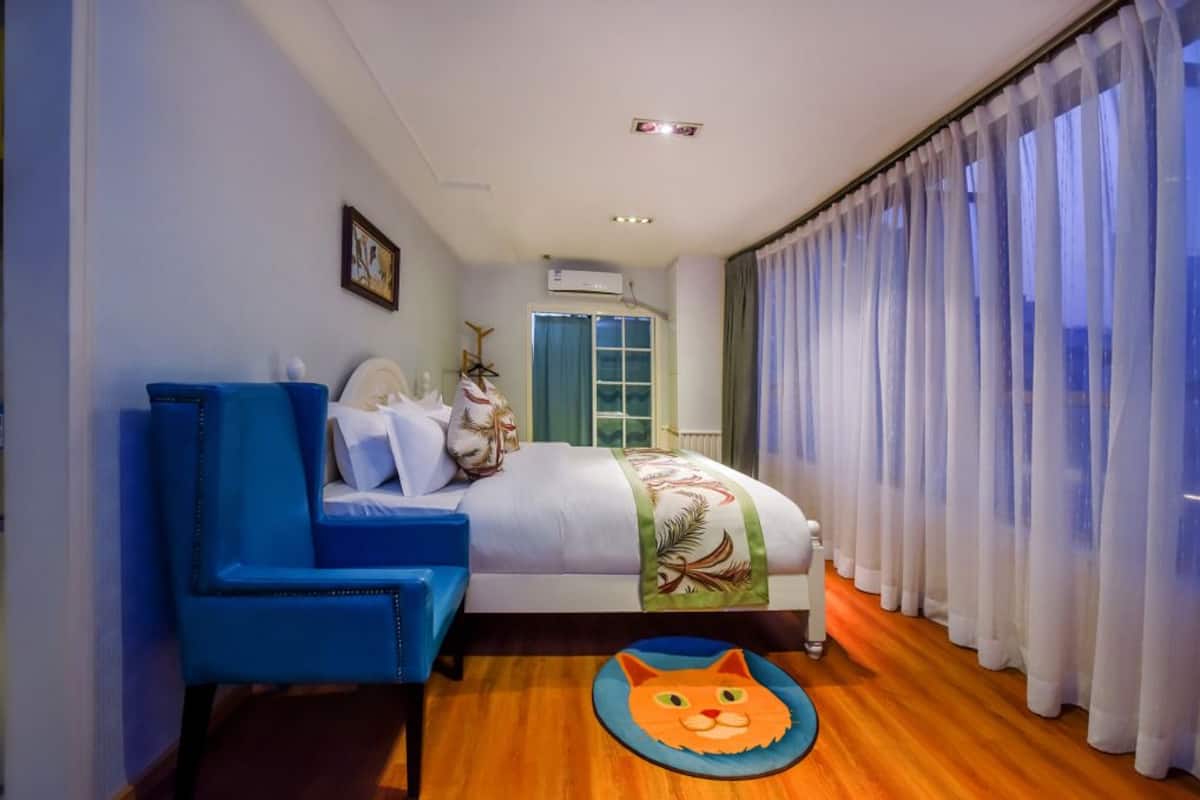 Quarto com cama, uma cadeira azul, janela com cortinas e um tapete em formato de gato no chão.