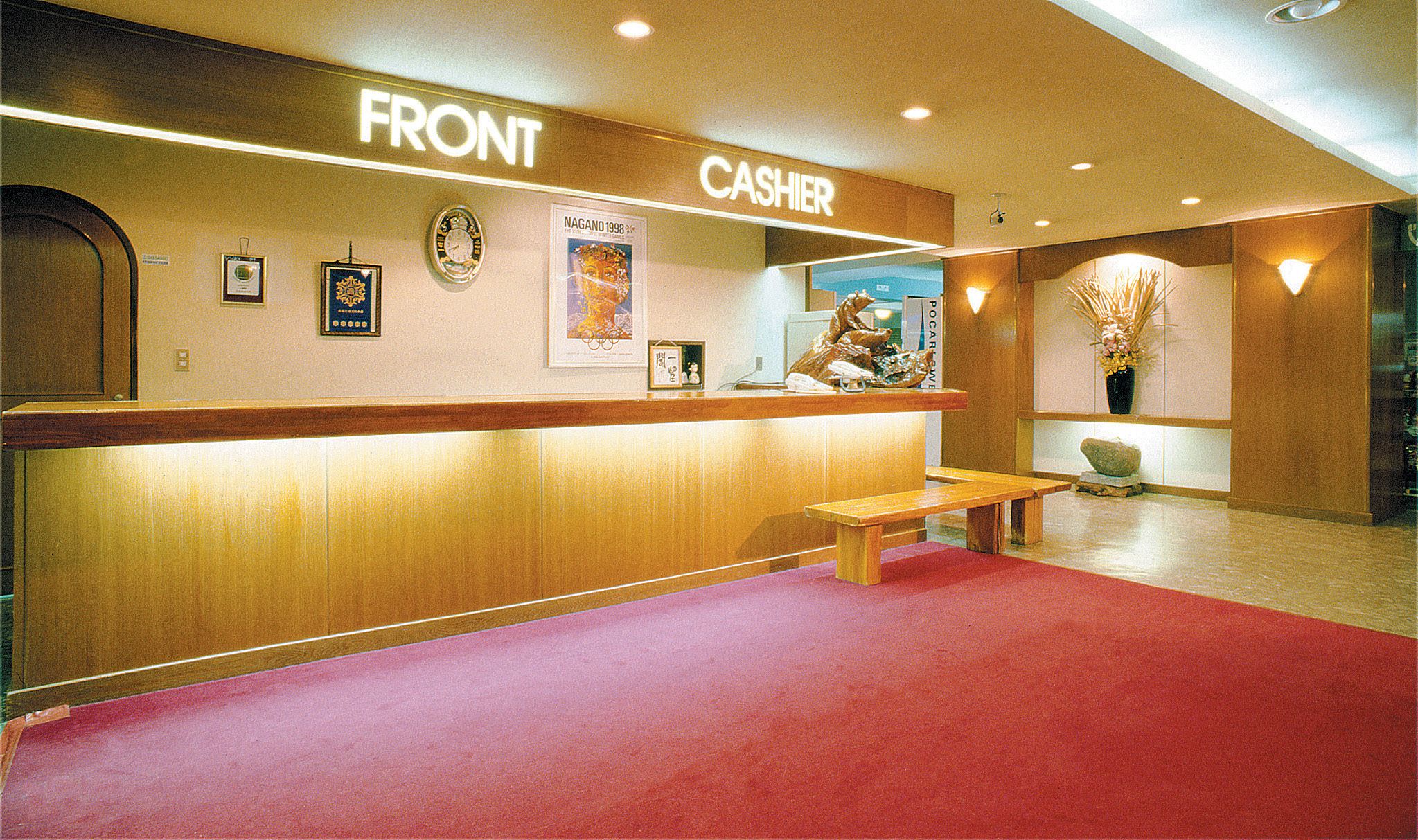 A hotel reception area with a wooden counter labeled "FRONT" and "CASHIER," a clock, framed pictures, a bench, and a red carpet.
