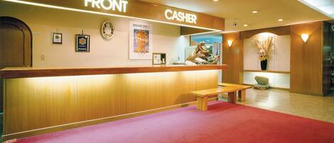 A hotel reception area with a wooden counter labeled "FRONT" and "CASHIER," a clock, framed pictures, a bench, and a red carpet.