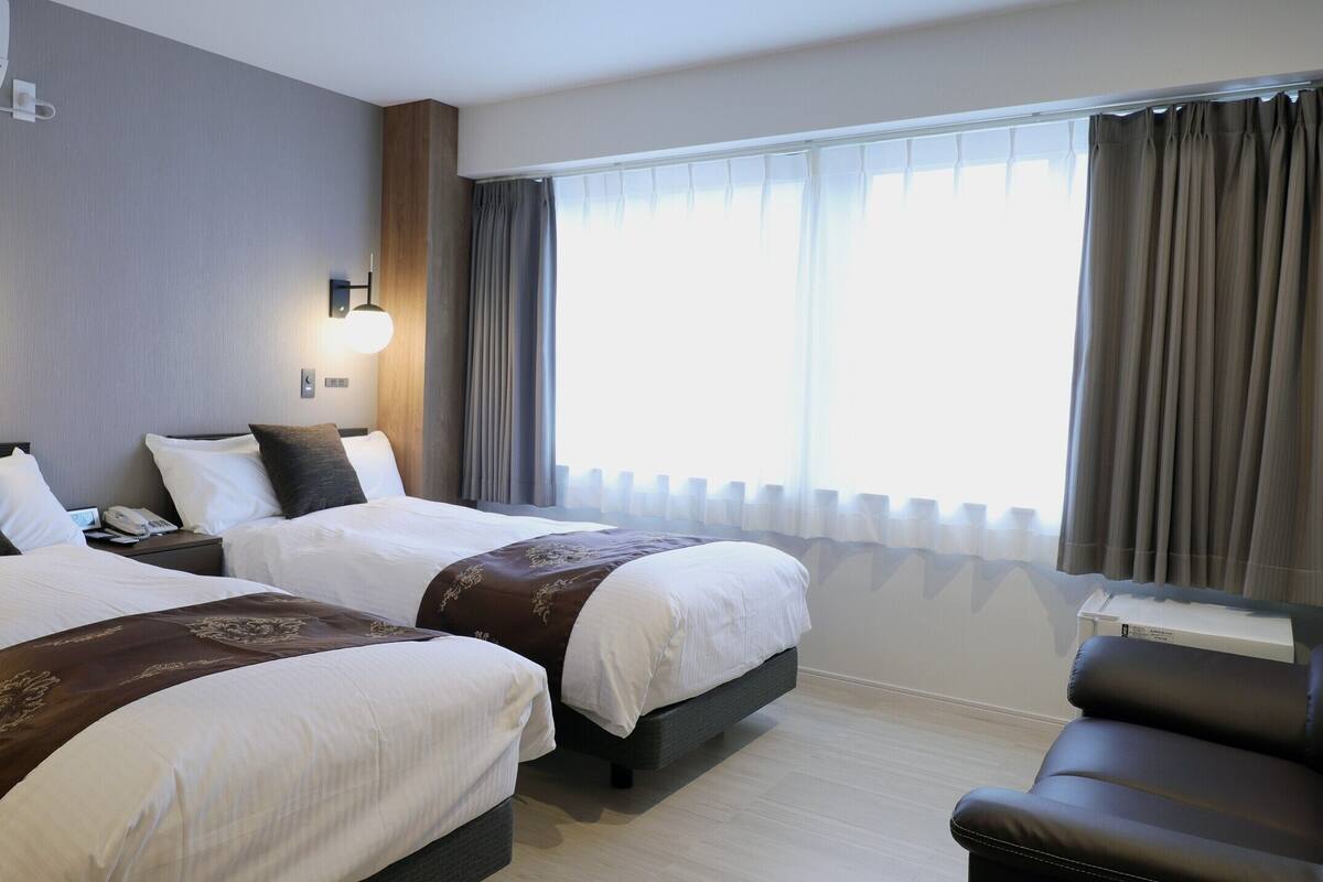 Habitació d'hotel amb dos llits, un sofà, una taula de nit i un telèfon, i una finestra amb cortines.