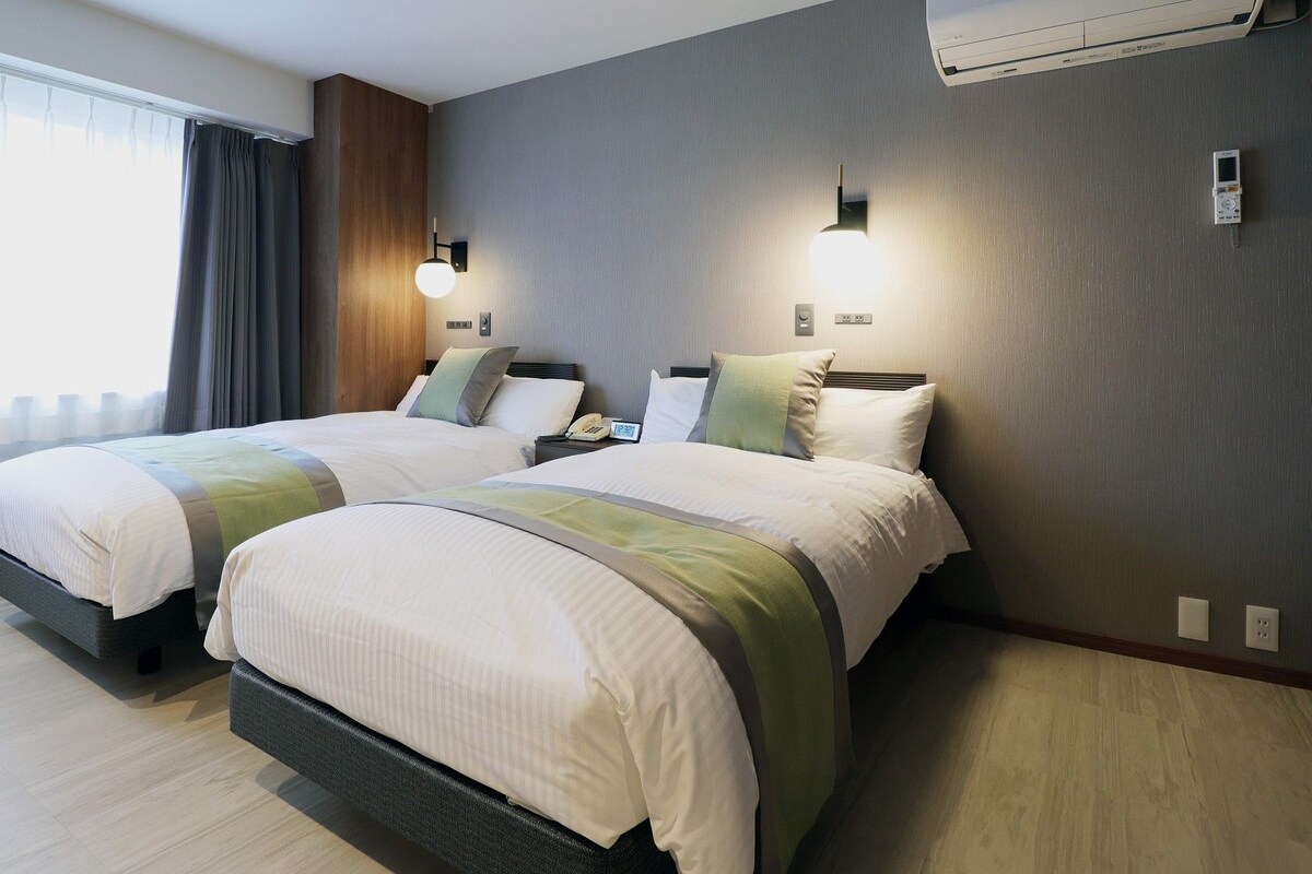 Une chambre d’hôtel avec deux lits, une fenêtre avec des rideaux, un climatiseur fixé au mur et une table de chevet avec un téléphone.