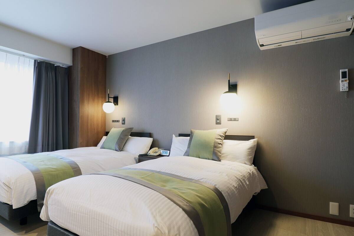 Habitació d'hotel amb dos llits, una finestra gran amb cortines, un aire condicionat fixat a la paret i una taula de nit amb una làmpada.