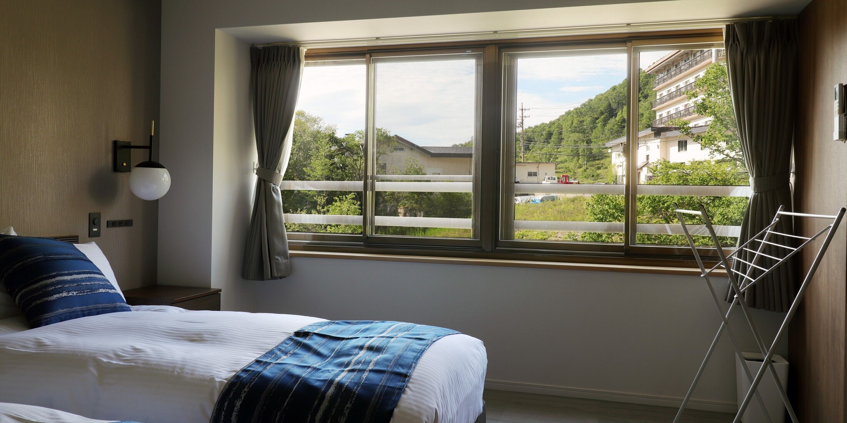 Habitación de hotel con una cama, una ventana que ofrece vista a una zona residencial y una lámpara fijada a la pared.