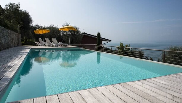 Ein rechteckiger Swimmingpool mit klarem, blauem Wasser, umgeben von einer Holzterrasse. Zwei gelbe Sonnenschirme spenden Schatten, und der Pool grenzt an ein Gebäude mit Blick auf das Meer.