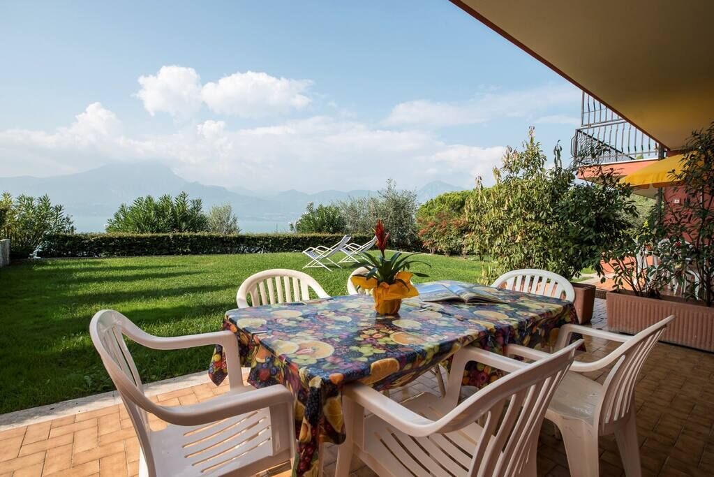 Een terras met een tafel en stoelen, uitzicht op een grasveld en bergen, en een gebouw met een balkon op de achtergrond.