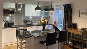 A modern kitchen with white cabinets, a central island with chairs, a microwave, a coffee maker, and a vase with flowers. - Kokopelli cottage near the Donkey sanctuary Sidmouth , modern and pet friendly. (Sidmouth)