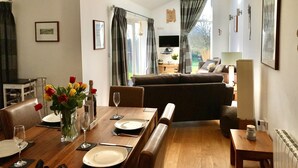 A dining area with a wooden table set for four, a sofa in the background, and a view of the outdoors through large windows. - Kokopelli cottage near the Donkey sanctuary Sidmouth , modern and pet friendly. (Sidmouth)