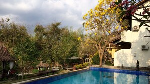 Una piscina con agua cristalina azul, rodeada de exuberante vegetación y un edificio blanco con tejado de baldosas.