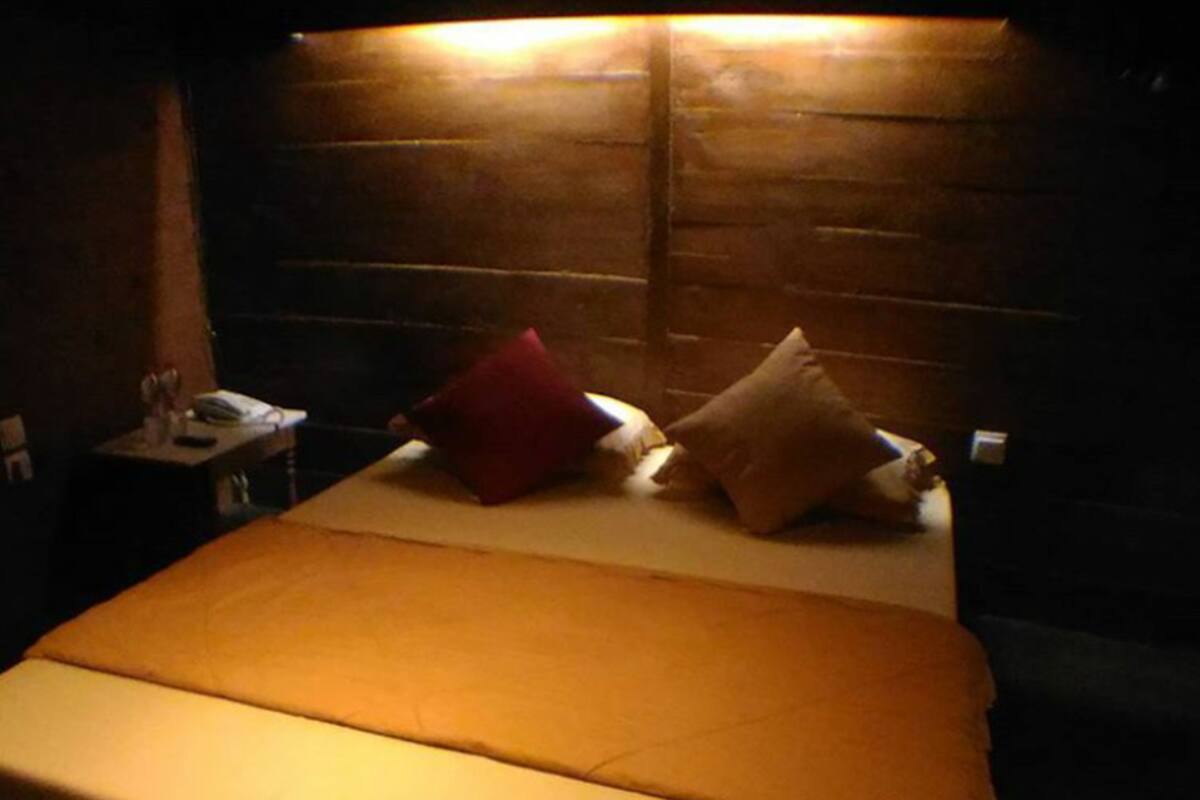 Quarto de hotel com cabeceira de madeira, cama com colchão branco e duas almofadas, e uma pequena mesa de cabeceira com abajur.