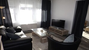 A modern living room with a sofa, a glass coffee table, a TV on a wooden stand, and a window with curtains. - Bright and modern apartment right on the North Sea (Nordstrand)